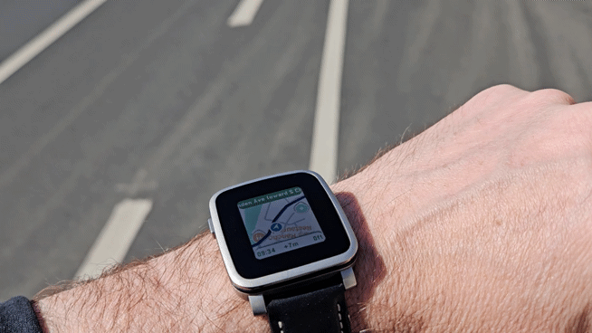 A Pebble Time Steel on a cyclist's wrist showing MirrorMap navigating in real time — a live map with the route drawn on it, the next-turn instruction, ETA and distance